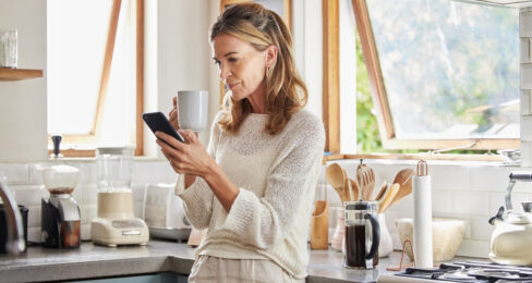 A middle age woman in her kitchen, drinking coffee and reading from her iPhone.