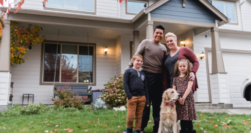 A couple standing outside of their home in the fall with their two young children and a golden retriever.
