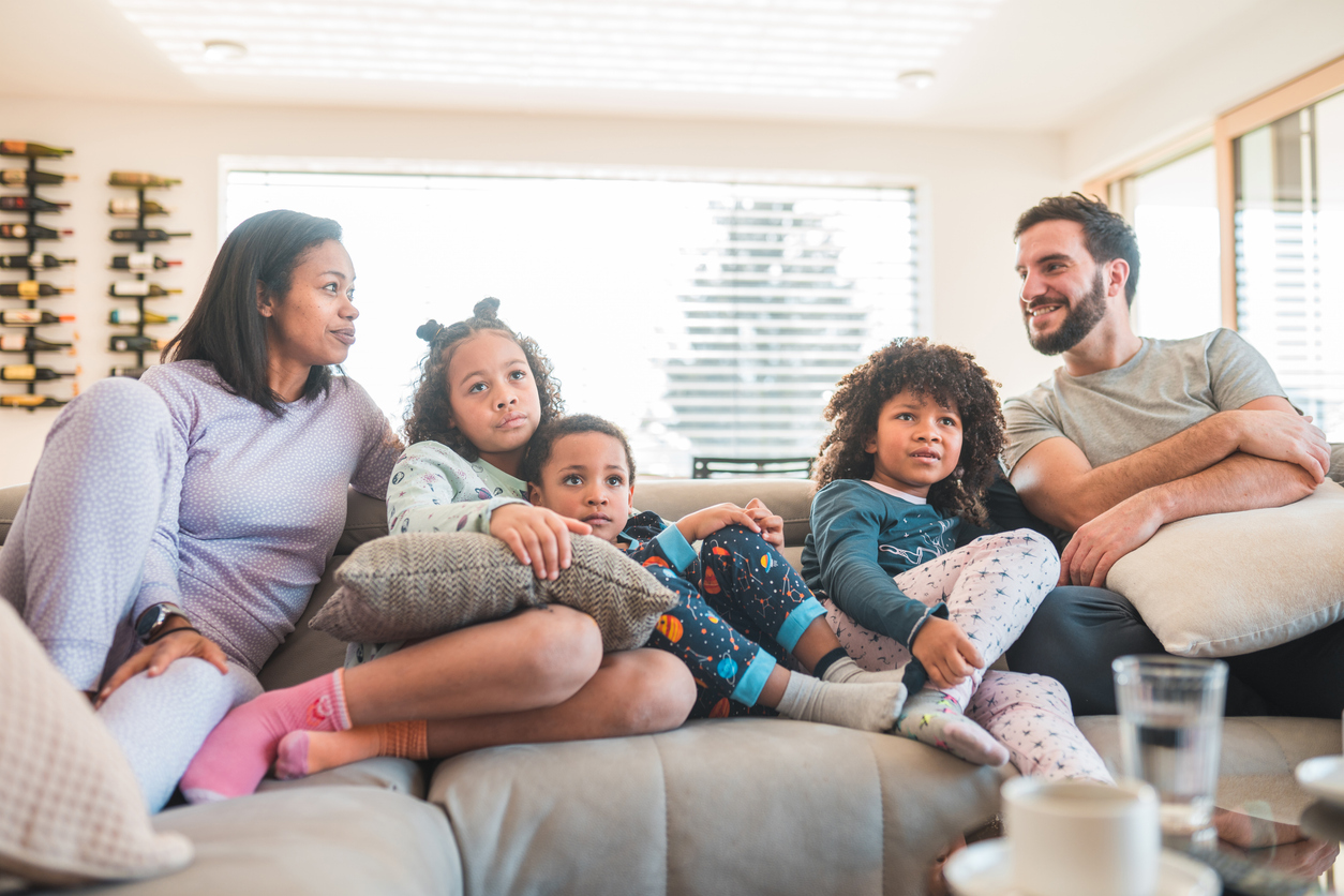 A couple and their three young children relax on the couch while the children watch tv.