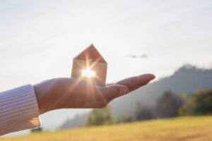 A woman's hand holds a small wooden house, with the sun shining behind it.